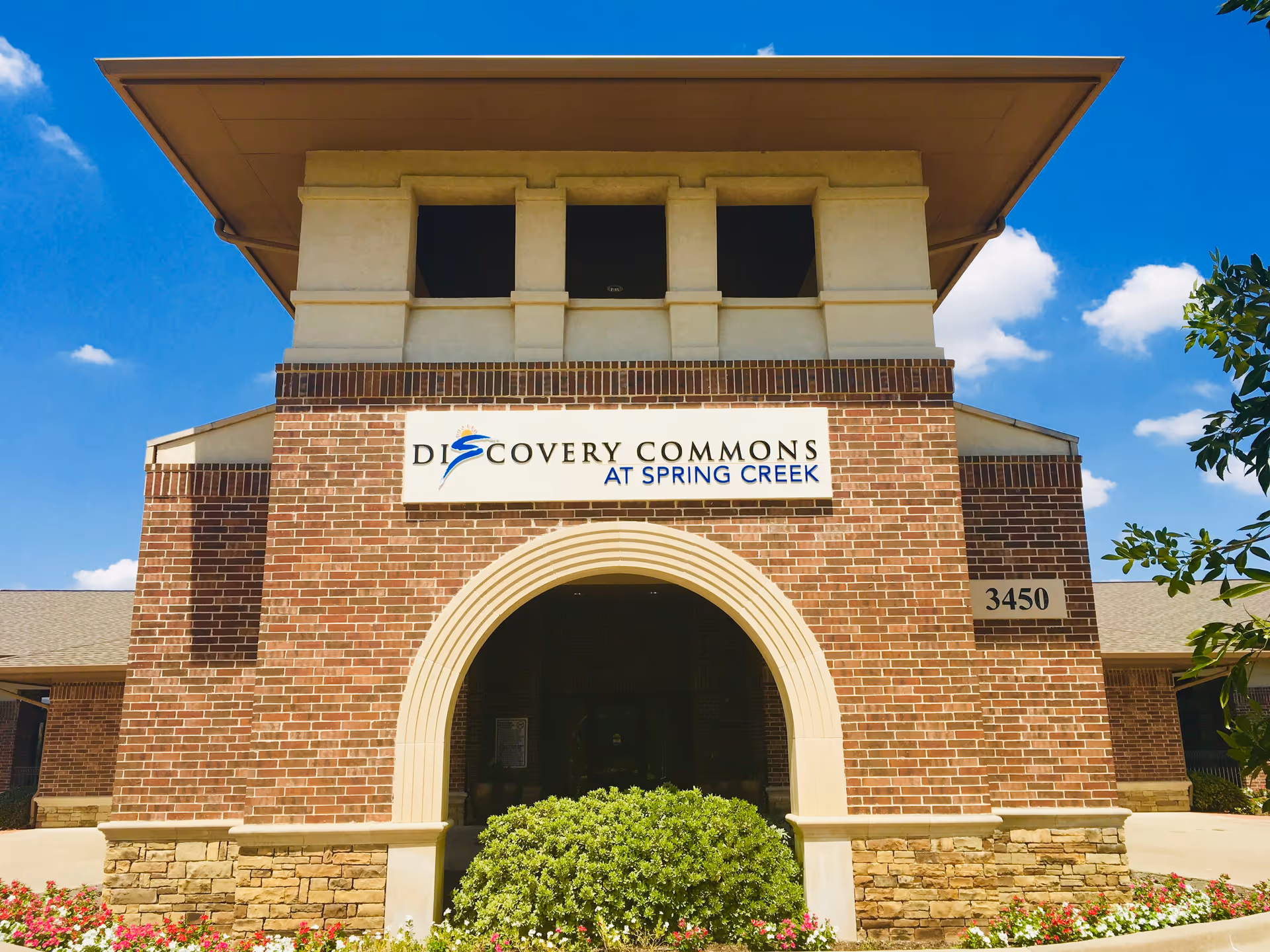 Front exterior view of a brick building with a large archway entrance. Above the archway is a sign that reads 'Discovery Commons at Spring Creek'. The building number 3450 is displayed on the right side. There are bushes and flowers in front of the building under a clear blue sky with some clouds.