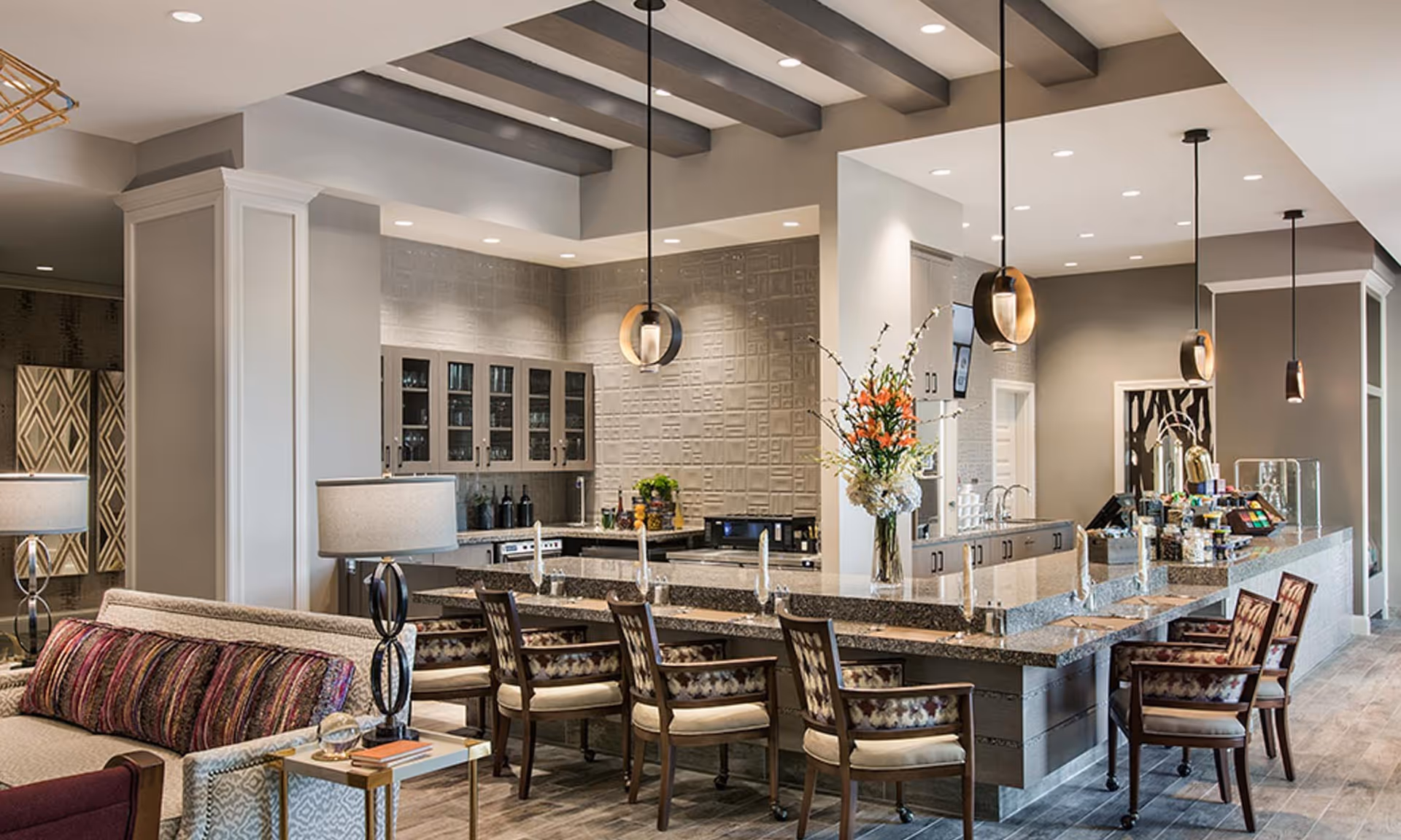 A modern and spacious interior area featuring a large kitchen island with granite countertops surrounded by several wooden chairs with patterned cushions. The kitchen area has glass-front cabinets, a textured backsplash, and pendant lights hanging from the ceiling. In the foreground, there is a cozy seating area with a sofa adorned with colorful pillows and a side table with a lamp and books. A vase with flowers is placed on the kitchen island, adding a decorative touch.