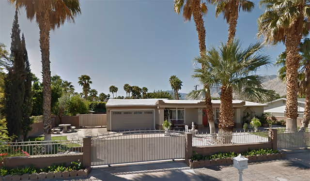Single-story gated house with a two-car garage, palm trees in the yard, and mountains in the background.