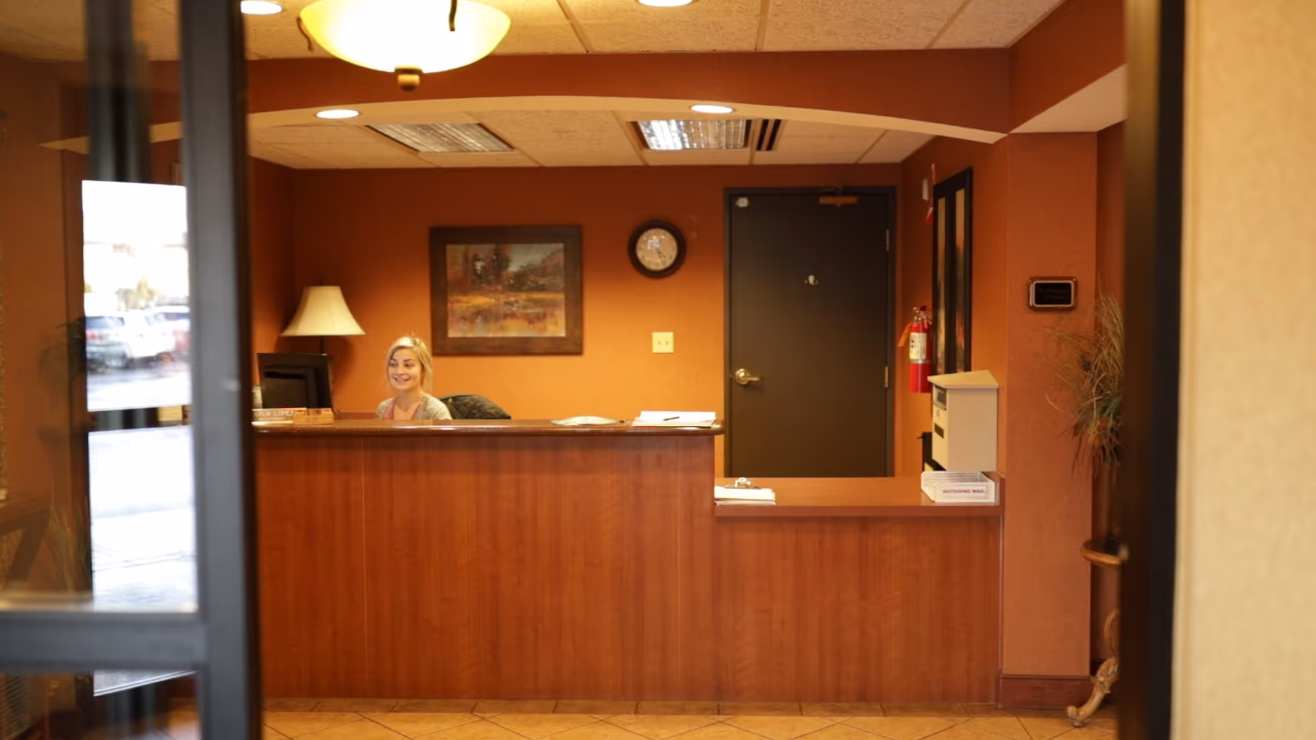 Reception area with a wooden front desk, a smiling receptionist seated behind the desk, a lamp on the left side, a framed painting and a clock on the wall, and a closed dark door in the background.