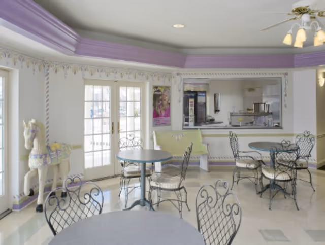 Bright indoor dining area with round tables and wrought-iron chairs, pastel purple trim, and a serving window.