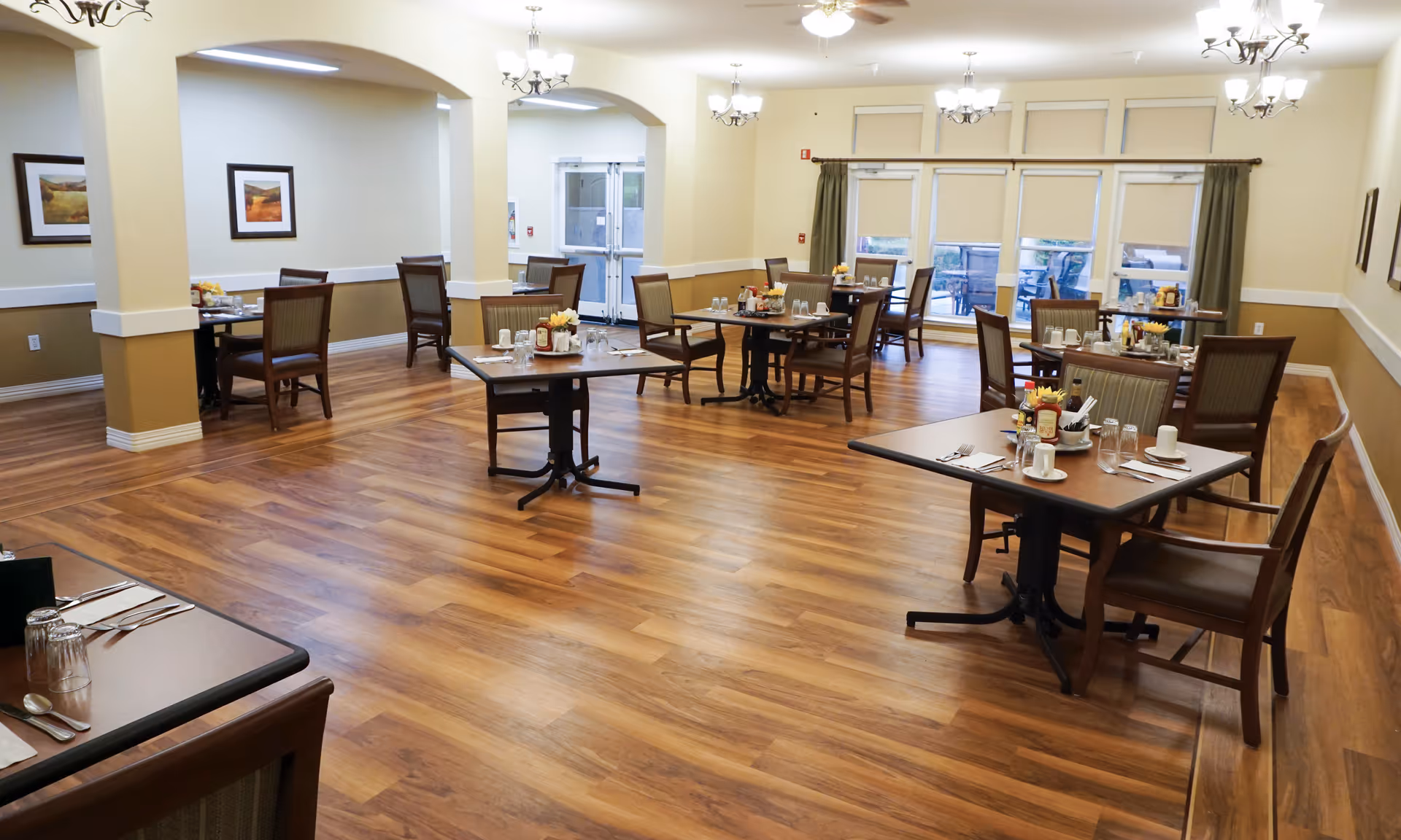 Spacious dining room with set tables and chairs on wood floors, chandeliers, and large windows.