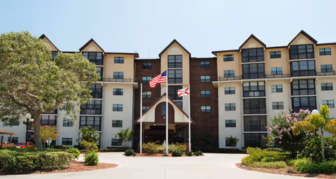Front exterior view of a multi-story senior living facility building with beige and brown walls, multiple windows, and peaked roof sections. Two flagpoles with the American flag and another flag stand in front of the entrance, surrounded by landscaped greenery and trees.