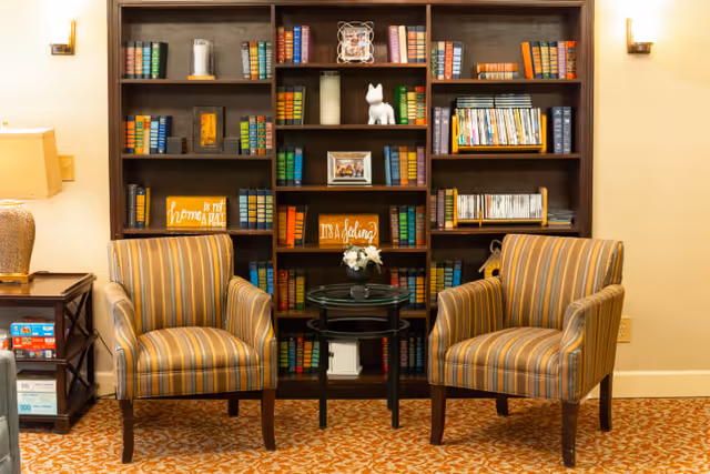 A cozy sitting area with two striped armchairs facing each other and a small round glass-top table between them. Behind the chairs is a large dark wooden bookshelf filled with colorful books, DVDs, decorative items, and framed pictures. The floor is covered with a patterned carpet, and a lamp is visible on a side table to the left.