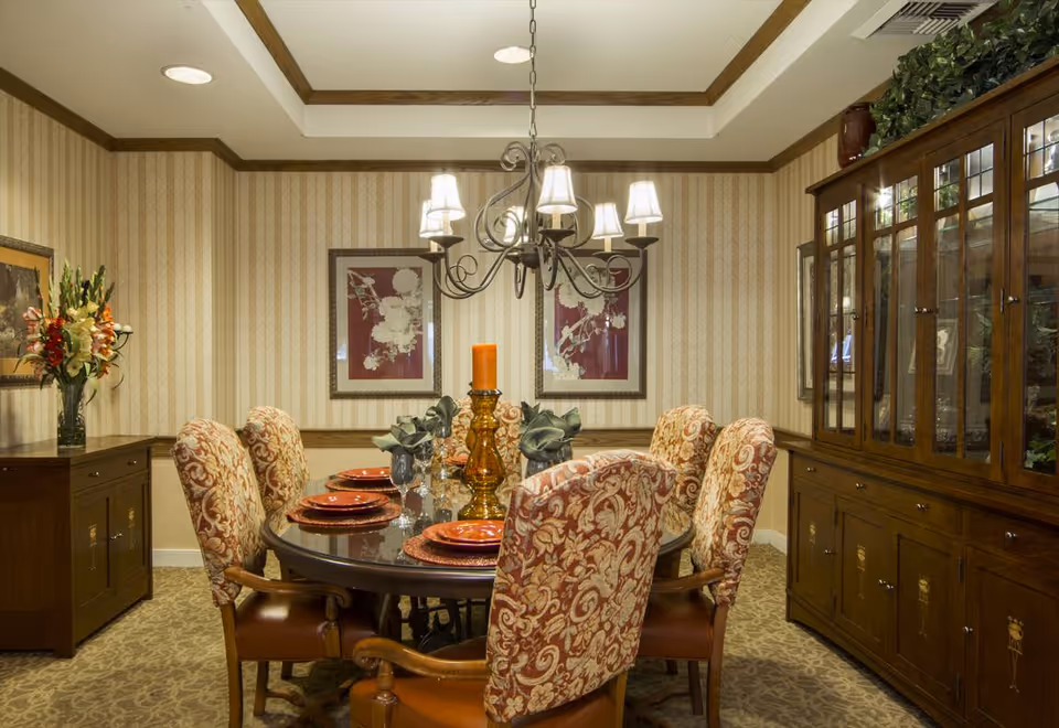 A formal dining room with a round wooden table set for six people. The chairs have ornate patterned upholstery. The table is decorated with orange plates, glassware, a large amber candle holder with an orange candle, and green napkins. The room features striped wallpaper, a chandelier with six lights, framed floral artwork on the wall, a wooden sideboard with a vase of flowers, and a large wooden china cabinet with glass doors displaying decorative items.