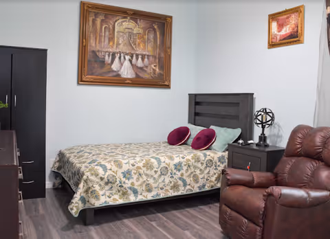 A bedroom with a bed covered in a floral patterned quilt, two round purple pillows, a dark wooden headboard, a matching nightstand with a decorative lamp, a brown leather recliner chair, a dark wardrobe, and a framed painting of dancers on the wall.