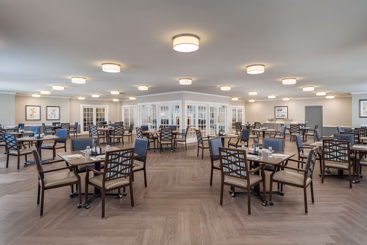 A spacious dining room in a senior living facility with multiple wooden tables and chairs arranged neatly. The room features light-colored walls, framed artwork, and numerous ceiling lights providing bright illumination. There is a central area with glass-paneled doors and windows, and the floor has a wood-patterned design.