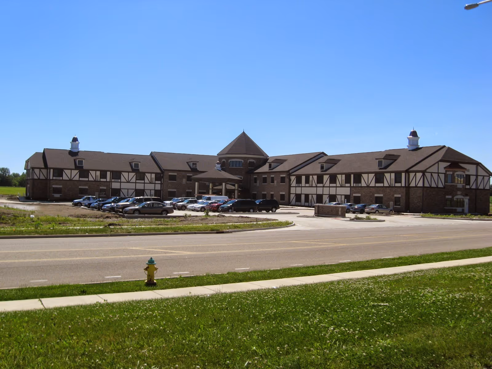 Exterior view of a large two-story supportive living facility named Castle Manor with a Tudor-style design, multiple parked cars in front, a clear blue sky, and a green lawn with a fire hydrant in the foreground.