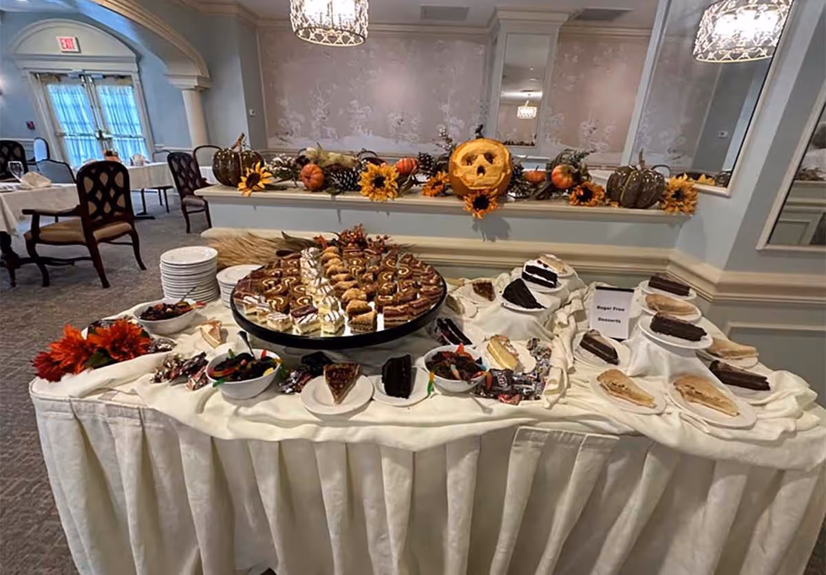 Buffet table of assorted cakes and pastries decorated with pumpkins and a jack-o'-lantern in a dining room.