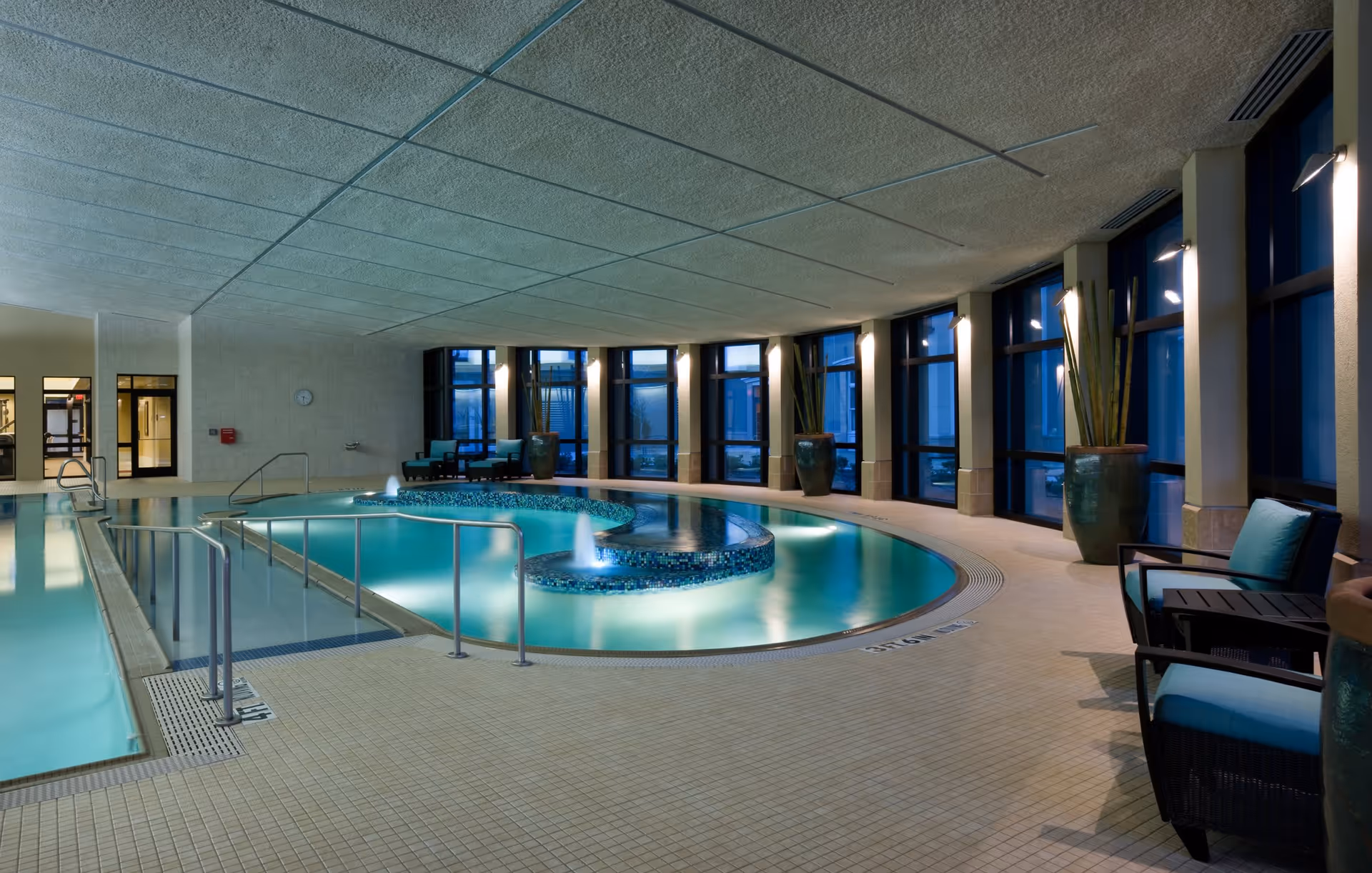 Indoor swimming pool area with a uniquely shaped pool featuring a tiled circular section with water fountains. The space has large windows along one side, several potted plants, cushioned chairs, and soft lighting creating a calm atmosphere.