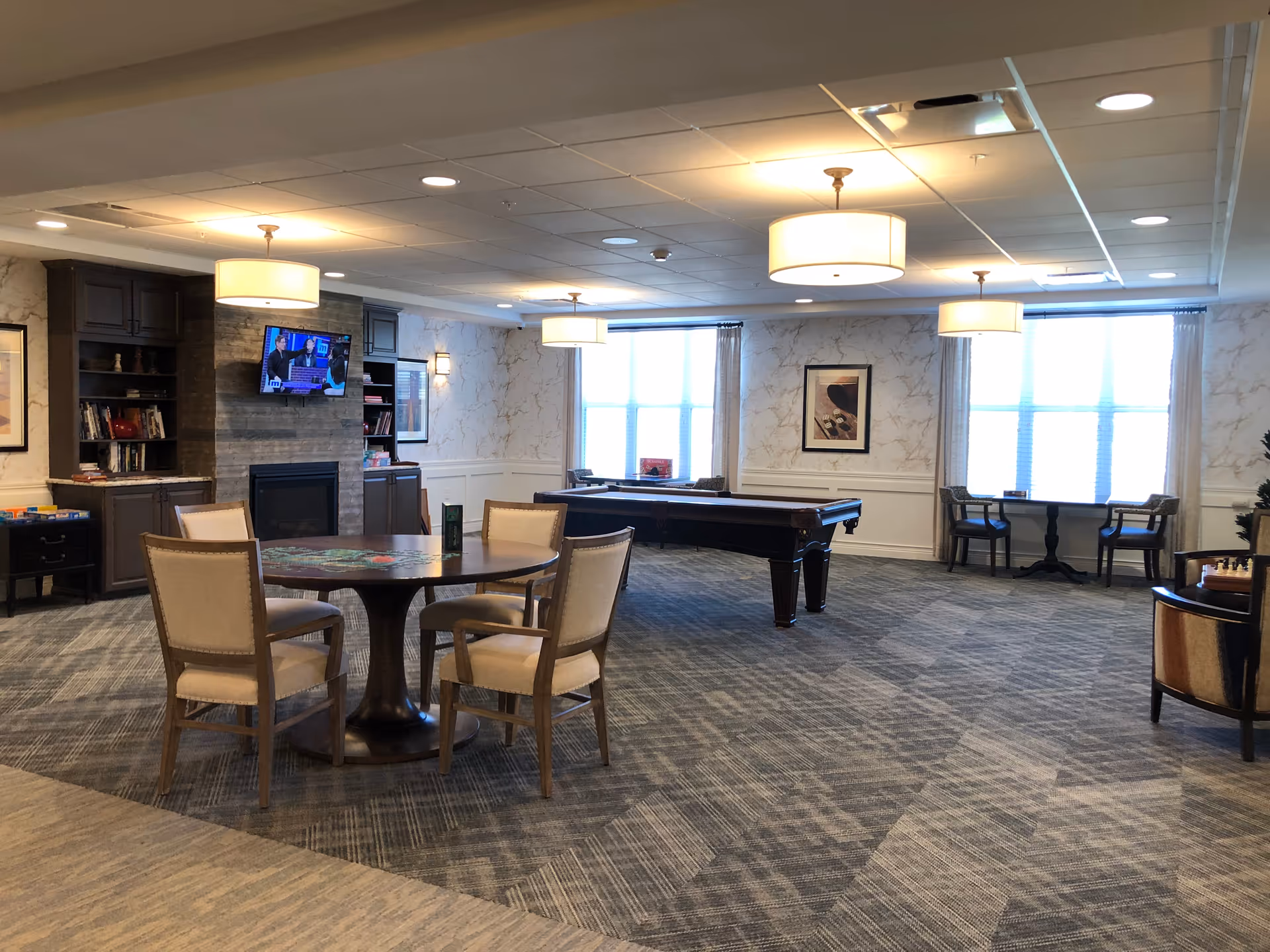 A spacious senior living common area with a round table and four chairs in the foreground, a pool table in the middle, and a fireplace with a mounted TV on the left wall. The room has large windows with curtains, framed artwork on the walls, and multiple ceiling lights providing warm illumination.
