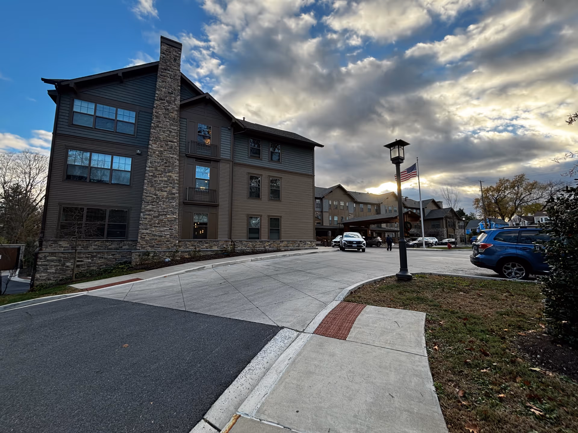 Exterior view of a multi-story senior living facility building with stone and wood siding under a cloudy sky. A driveway leads up to the entrance where cars are parked, and an American flag is flying on a flagpole near a streetlamp.