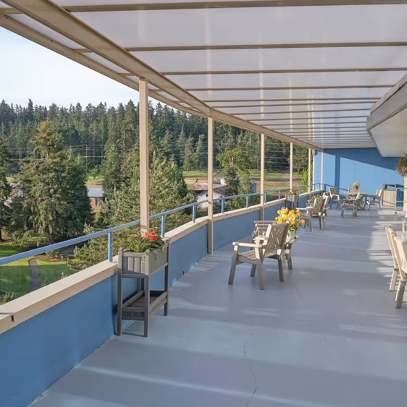 A covered outdoor balcony with chairs, potted flowers, and railings overlooking trees and a grassy area.
