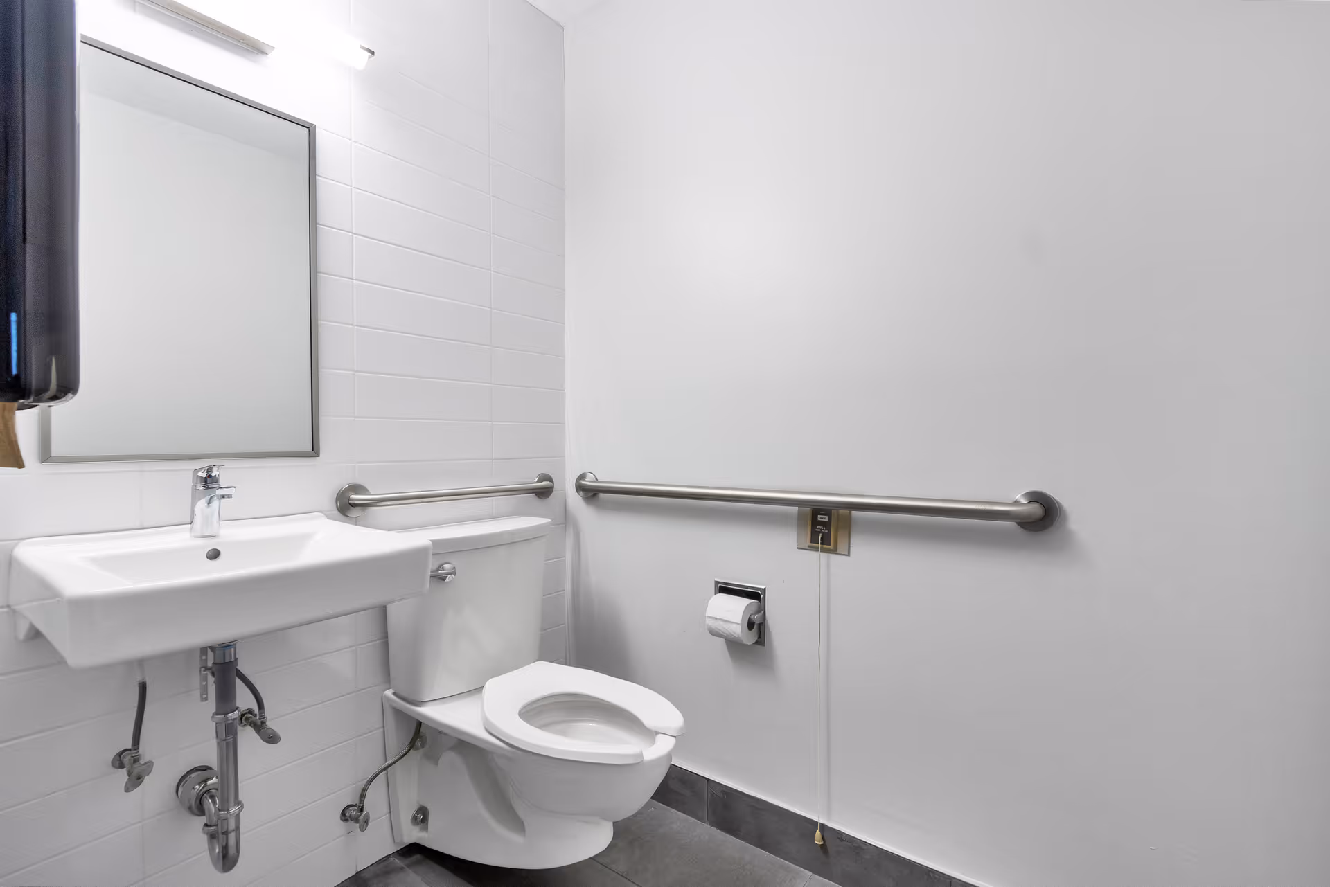 A clean and accessible bathroom with a white toilet, a wall-mounted white sink, a rectangular mirror above the sink, and stainless steel grab bars on the walls for support. There is a toilet paper holder with a roll of toilet paper and a pull cord on the wall next to the toilet. The walls are white and the floor is dark gray tile.