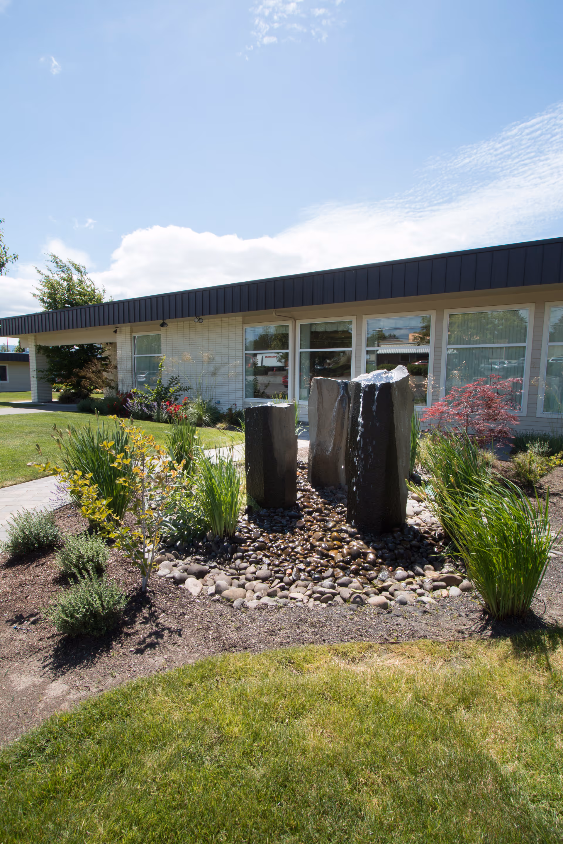 Front exterior of a single-story building with a three-stone water fountain and landscaped lawn under a blue sky.