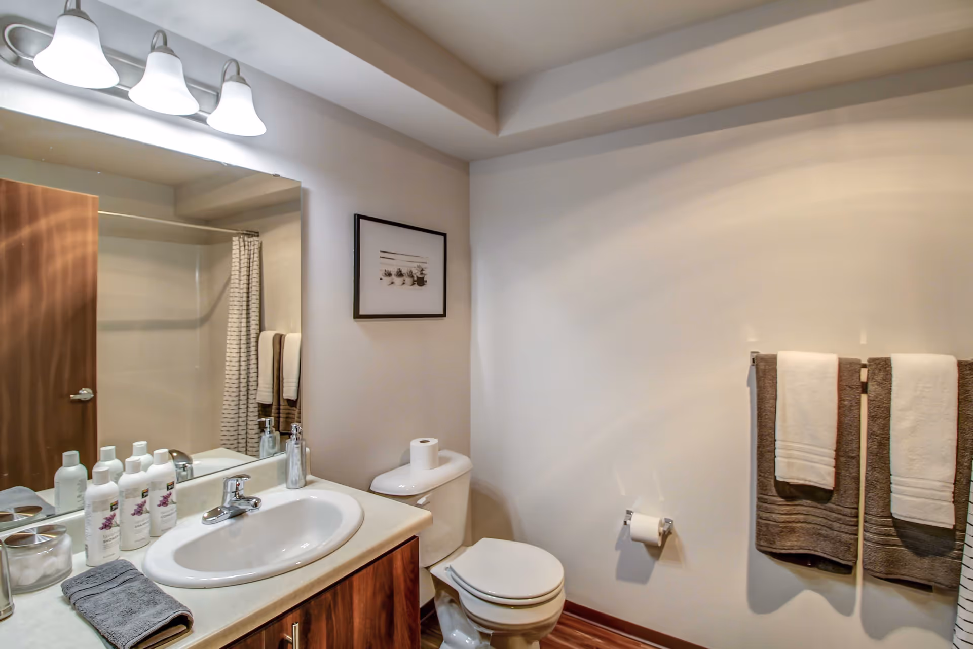 Well-lit bathroom with a sink and vanity, mirror, toilet, shower and towels hanging on a rack.