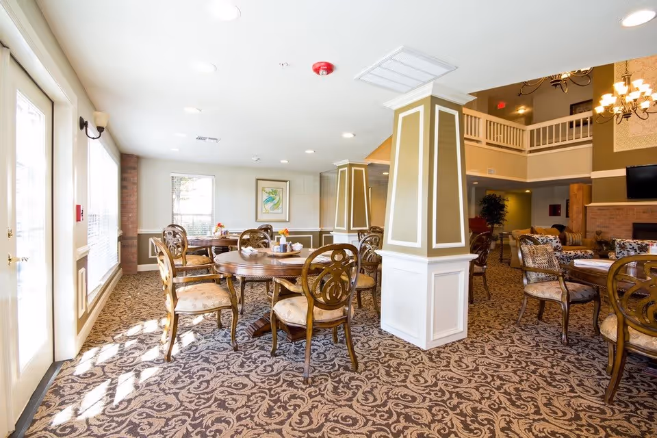 A bright and spacious common area with multiple round wooden tables and cushioned chairs arranged on a patterned carpet. The room features large windows allowing natural light to fill the space, decorative columns, a chandelier, and a fireplace with a mounted TV above it. The area has a warm and inviting atmosphere with neutral tones and tasteful decor.