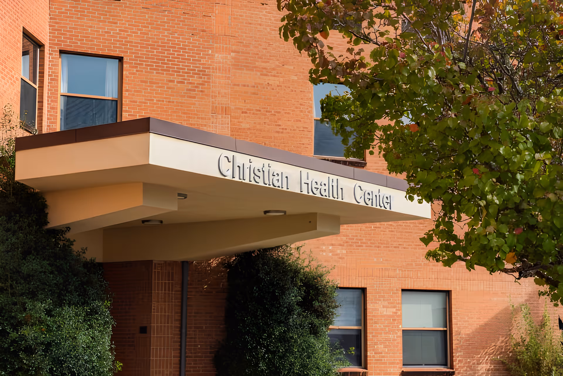 Entrance canopy of a brick building with the sign 'Christian Health Center' partially visible, surrounded by green bushes and a tree with green leaves.