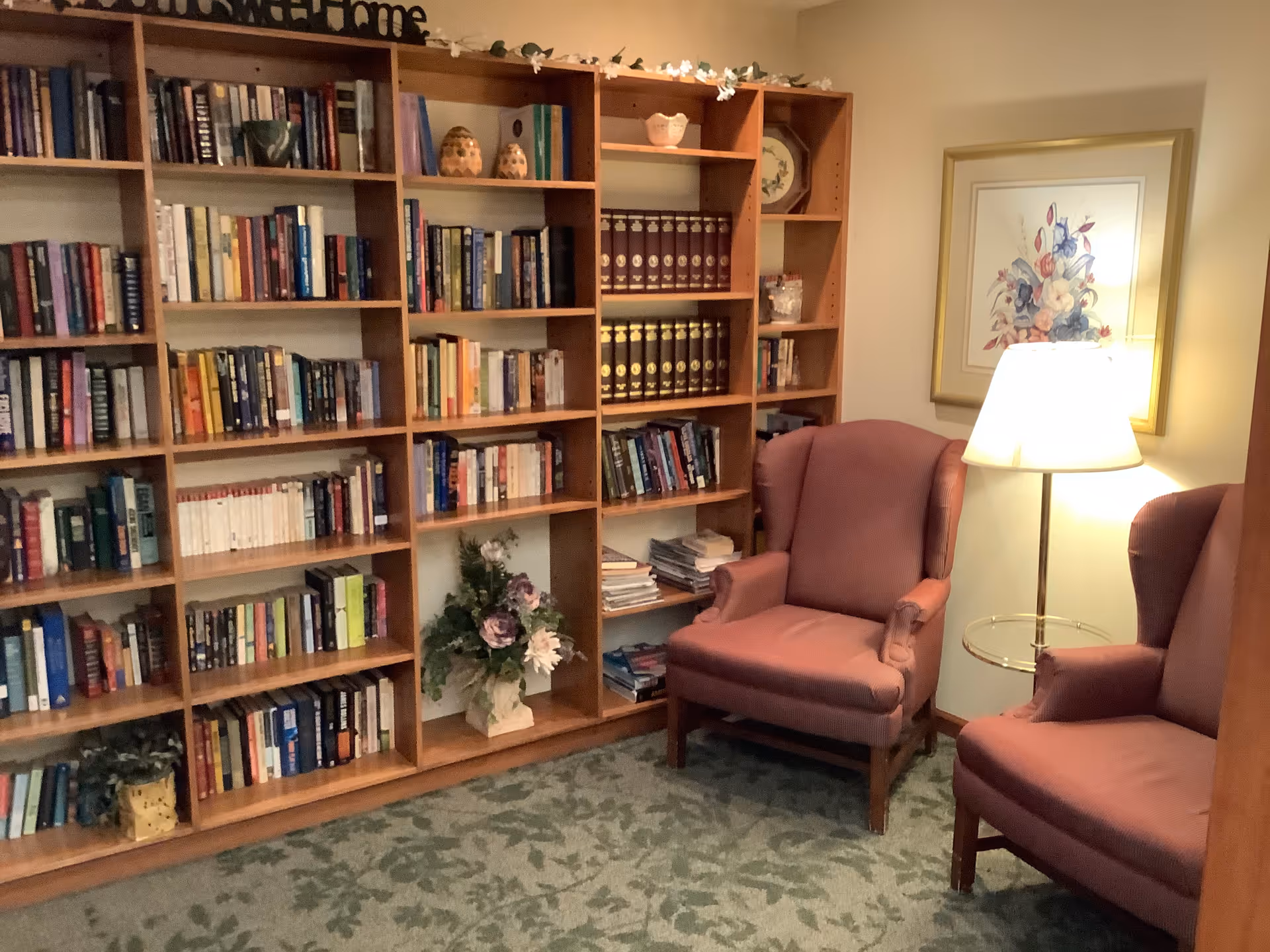 Small library-style sitting area with wooden bookshelves, two upholstered armchairs, a floor lamp, and floral decorations.