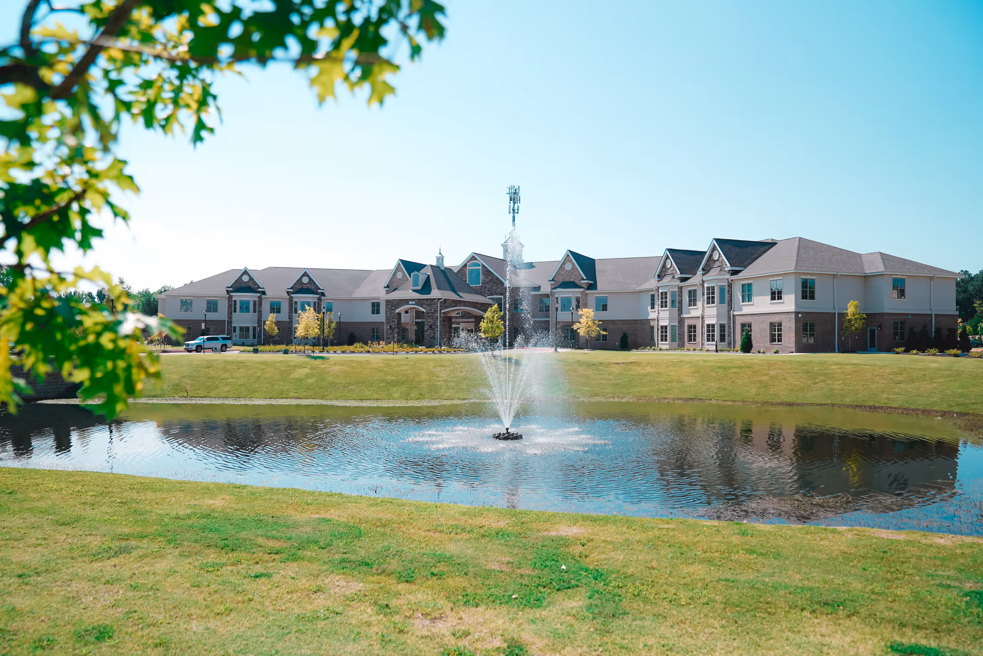 A large two-story building with a stone and beige exterior situated behind a pond with a water fountain in the center. The building is surrounded by a well-maintained grassy area with a few small trees, and the sky is clear and blue. Some green leaves from a nearby tree frame the left side of the image.