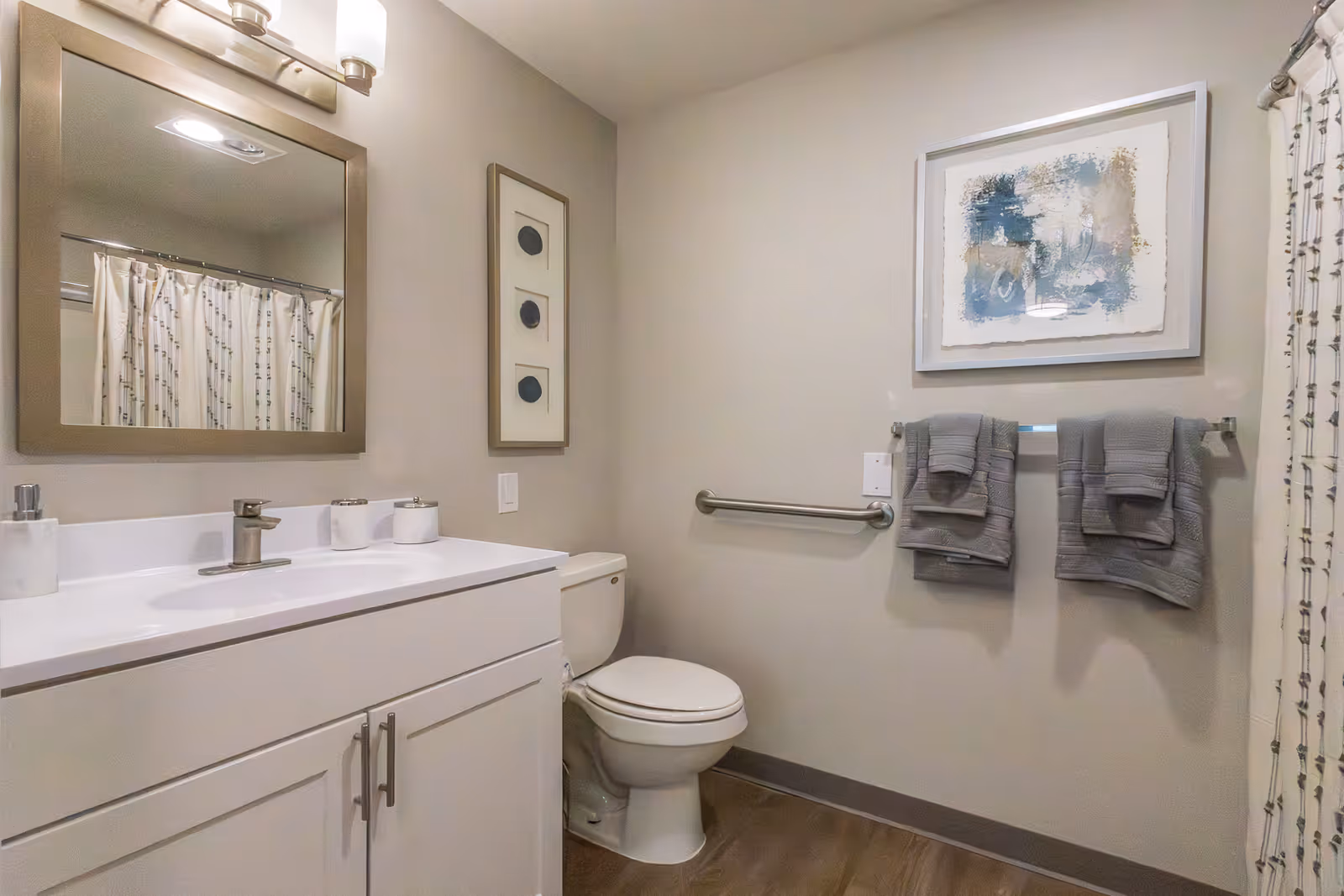 A clean and modern bathroom featuring a white vanity with a sink and faucet, a large mirror with three light fixtures above it, a toilet, a towel rack with two gray towels, a framed abstract painting on the wall, and a shower with a patterned curtain.
