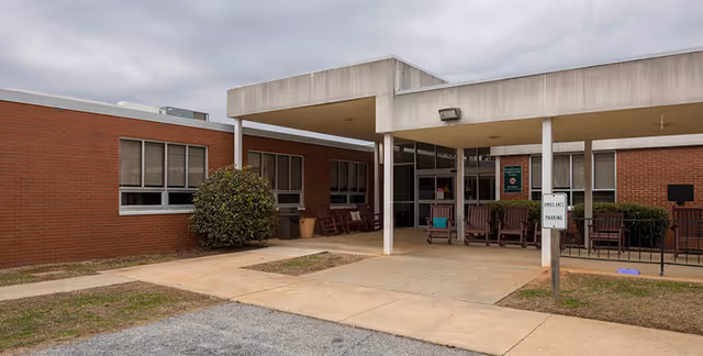 Exterior view of a single-story brick building with a covered entrance supported by white pillars. Several wooden benches are placed near the entrance, and a small sign indicates 'Emergency Parking.' The sky is overcast.