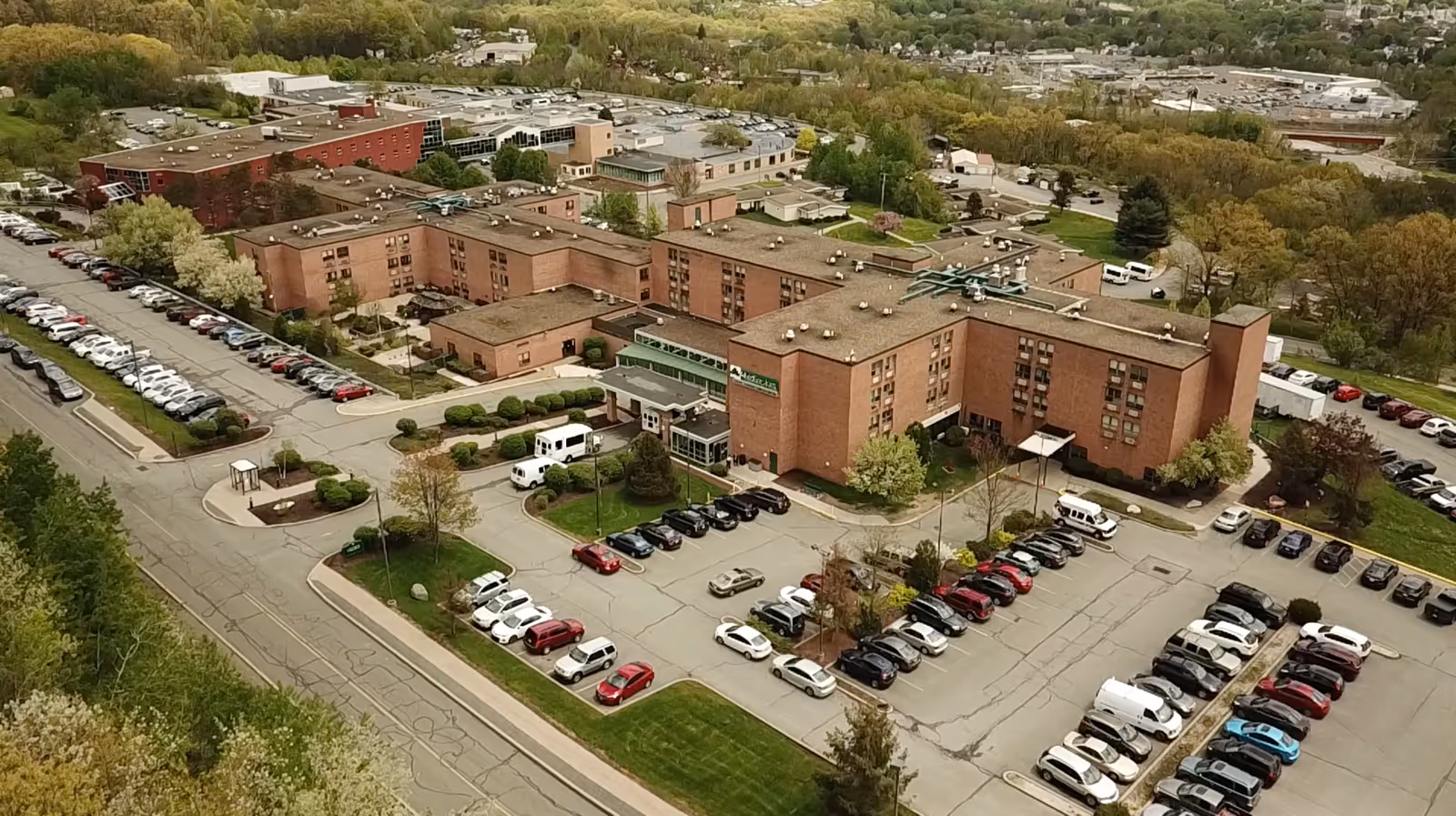Aerial view of a large multi-wing brick nursing facility surrounded by parking lots, cars, and trees.