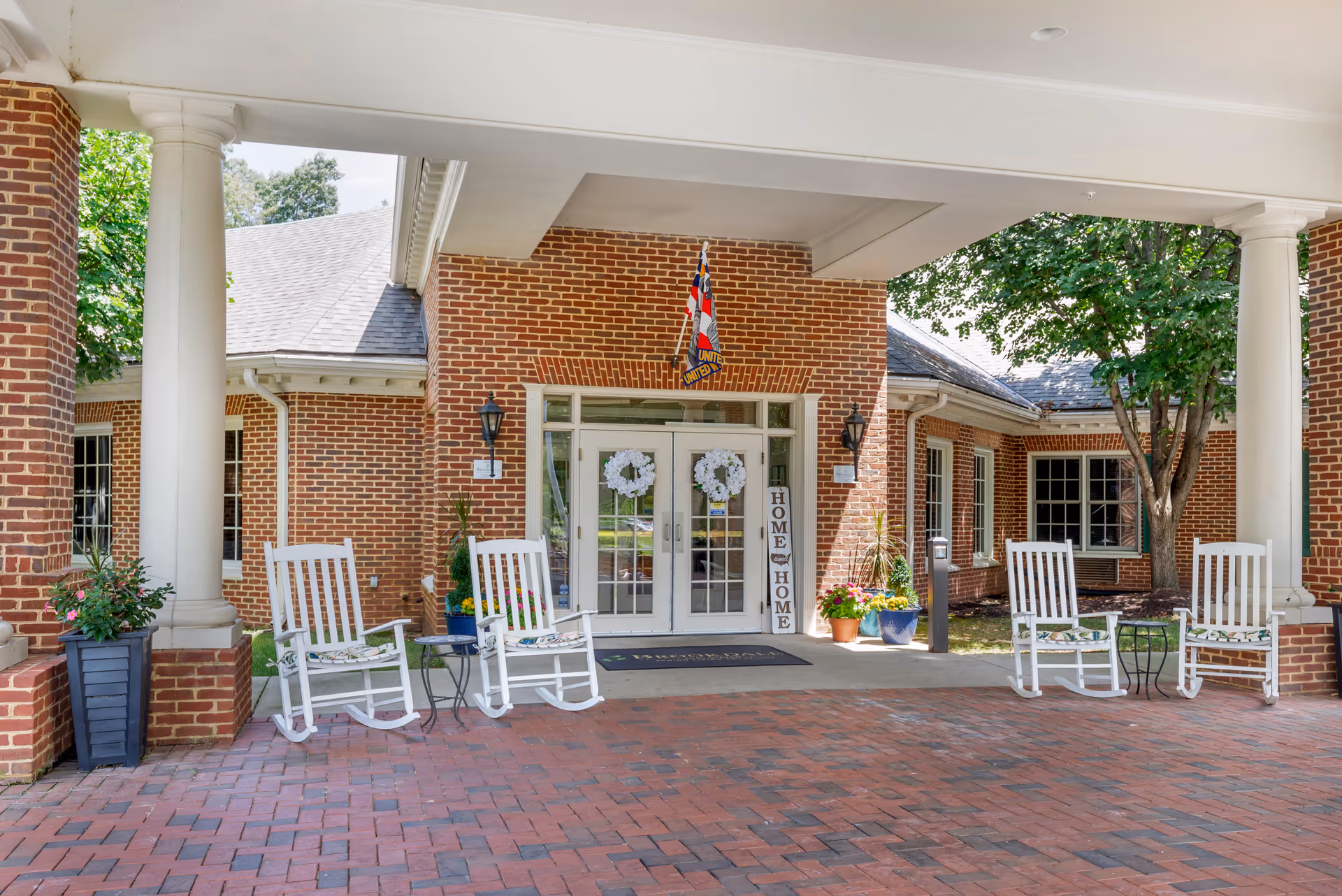 Entrance of a brick building with white double doors decorated with white wreaths. There are four white rocking chairs with floral cushions arranged on either side of the entrance, along with small tables and potted plants. A flag is hanging above the door, and a vertical sign next to the door reads 'HOME'.