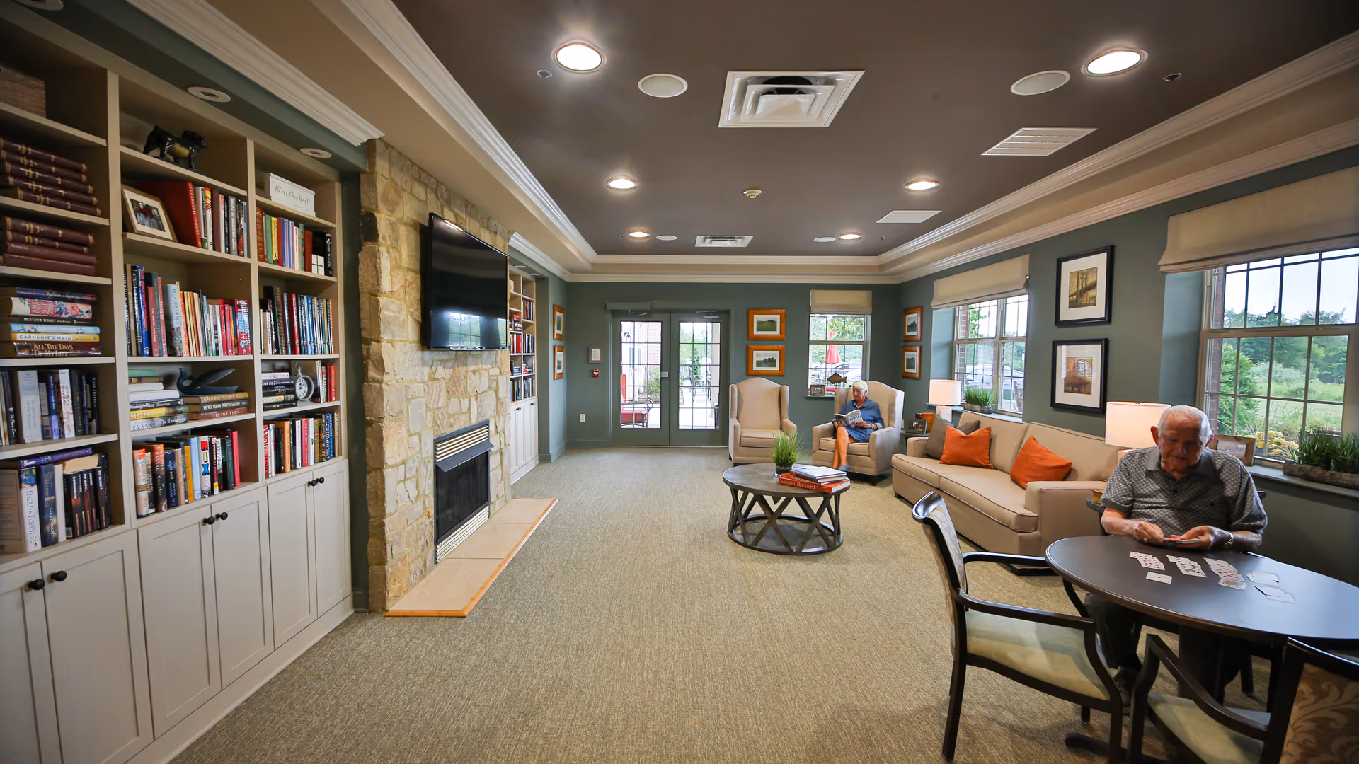 A cozy living room in Avalon Assisted Living at Hillsborough featuring built-in bookshelves filled with books, a stone fireplace with a mounted flat-screen TV above it, and comfortable seating including armchairs and a sofa with orange cushions. Two elderly residents are present; one is sitting on an armchair near the window reading, and the other is playing cards at a round table. The room has large windows letting in natural light and framed artwork on the walls.