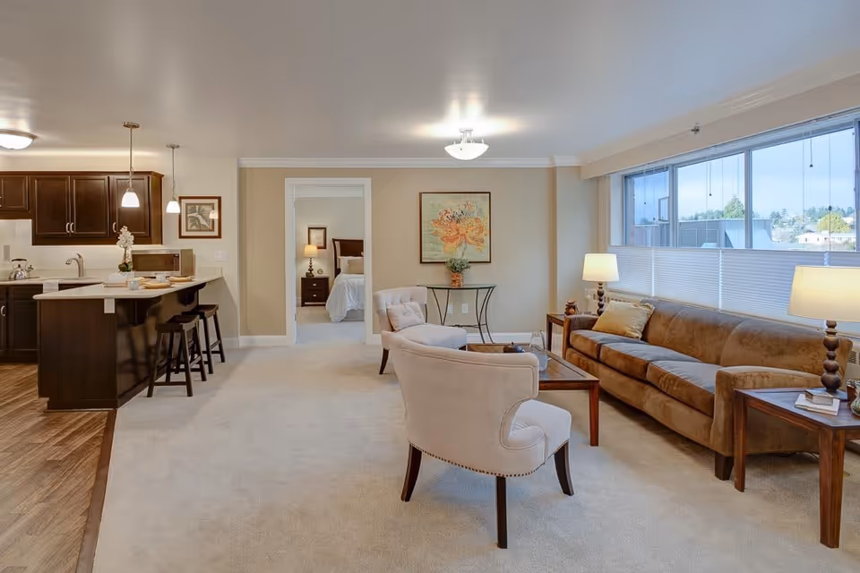 Open-plan living room and kitchen with a sofa, armchairs, coffee table, bar stools, and a large window.