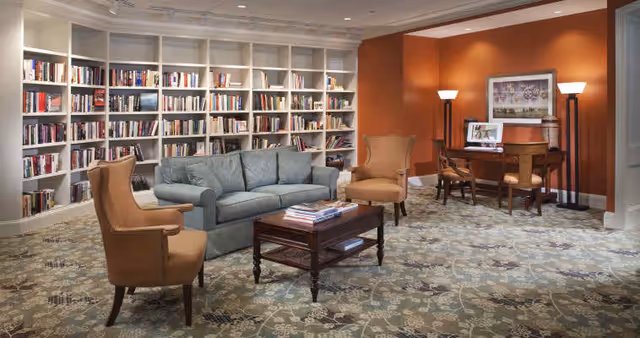 A cozy communal library-style sitting room with built-in bookshelves, a sofa and armchairs around a coffee table and a desk against an orange accent wall.