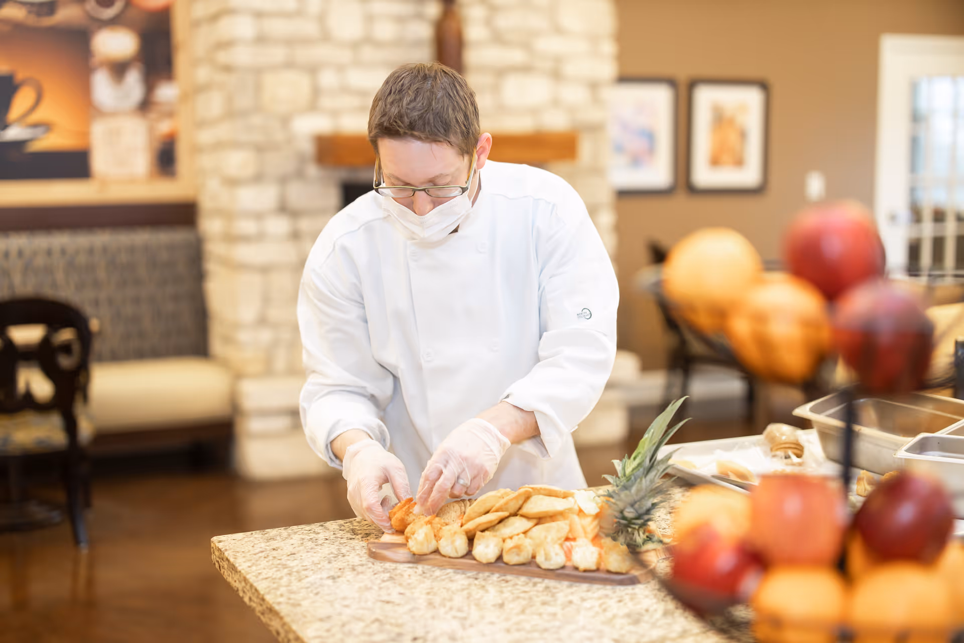 A chef wearing a white coat, gloves, and a face mask is preparing a platter of assorted pastries and bread on a kitchen counter in a warm, inviting dining area with stone walls and framed pictures.