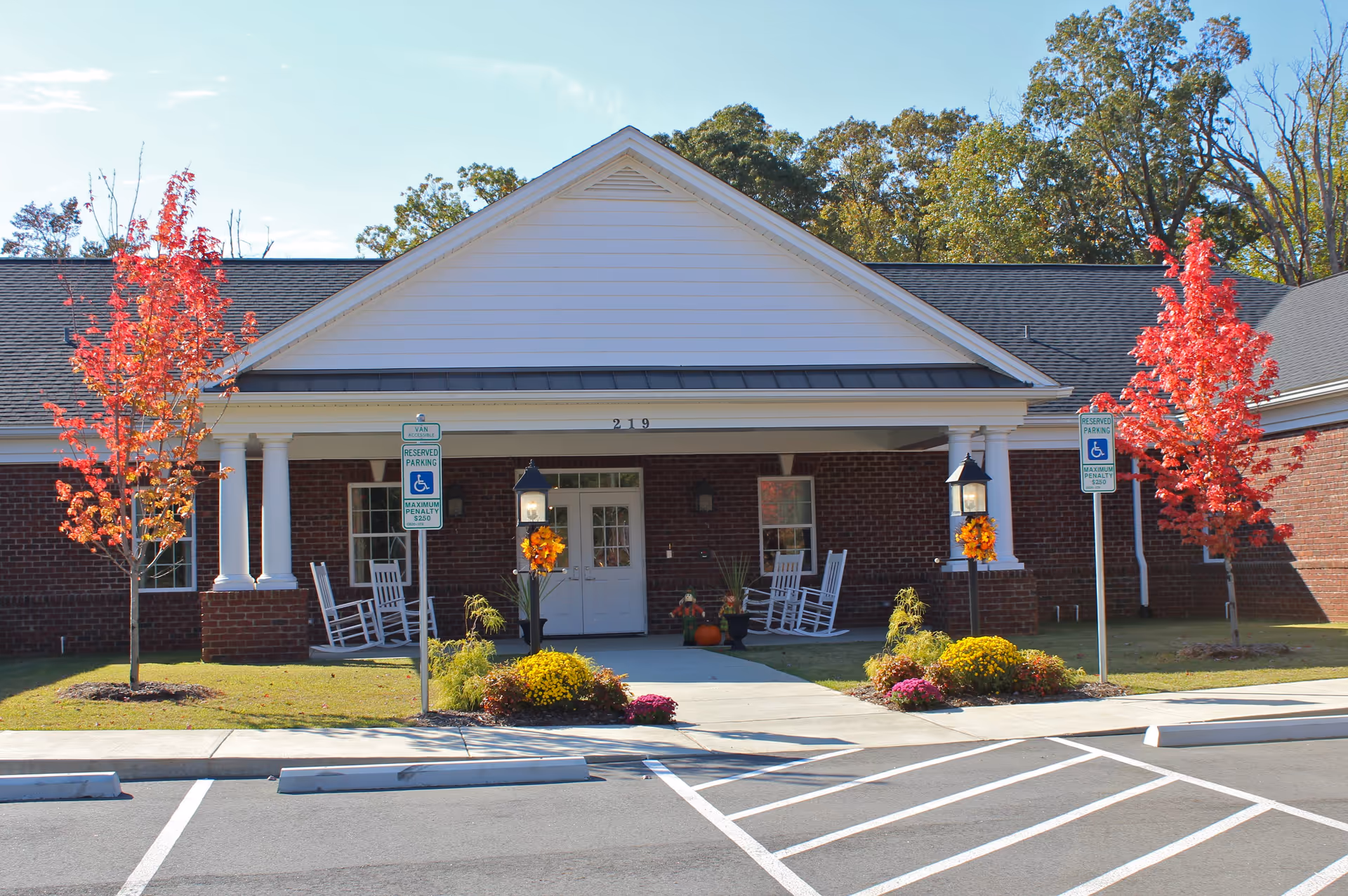 Front exterior view of a single-story brick building with a white gabled roof and columns at the entrance. There are two red-leaved trees on either side of the entrance, two reserved parking signs for handicapped parking, and two black lamp posts decorated with autumn wreaths. White rocking chairs are placed on the porch near the entrance door, which is flanked by windows. The building number 219 is displayed above the door.