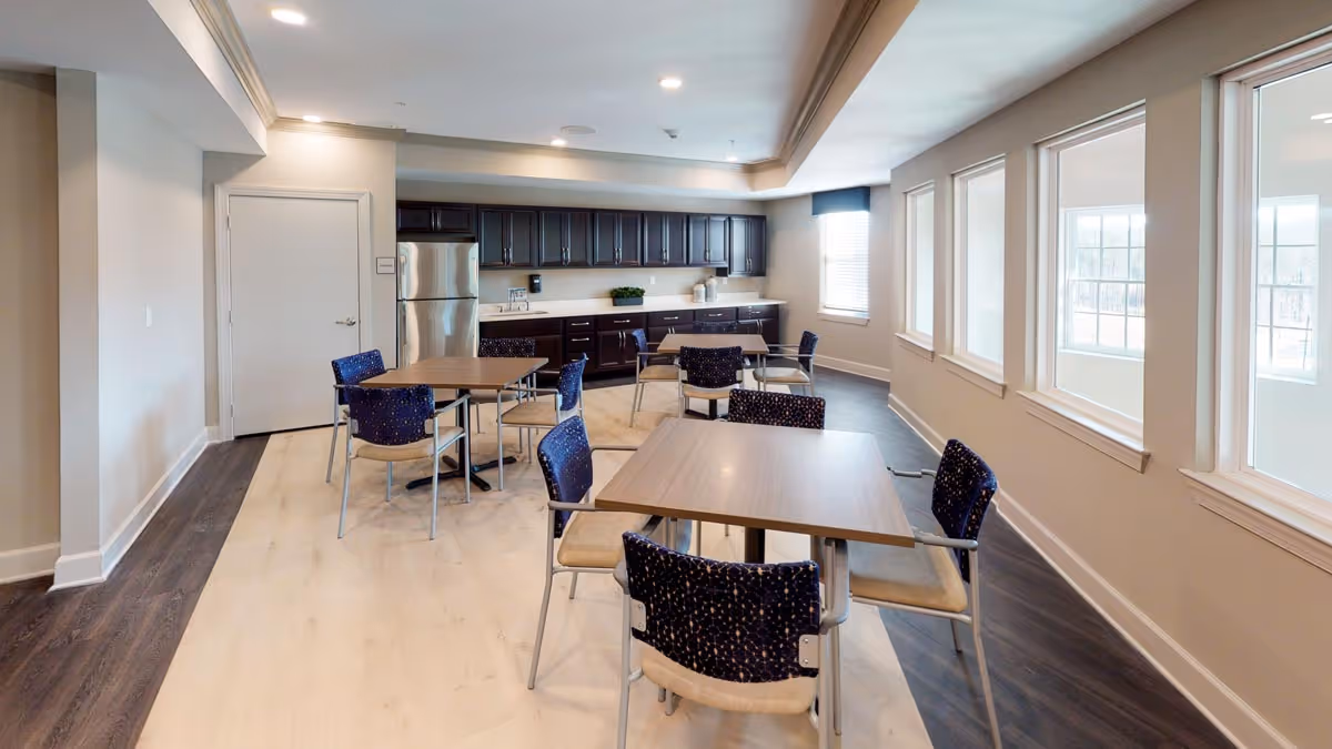 Communal dining area with several tables and chairs, a kitchenette with stainless steel refrigerator and dark cabinets, and a row of windows along one wall.