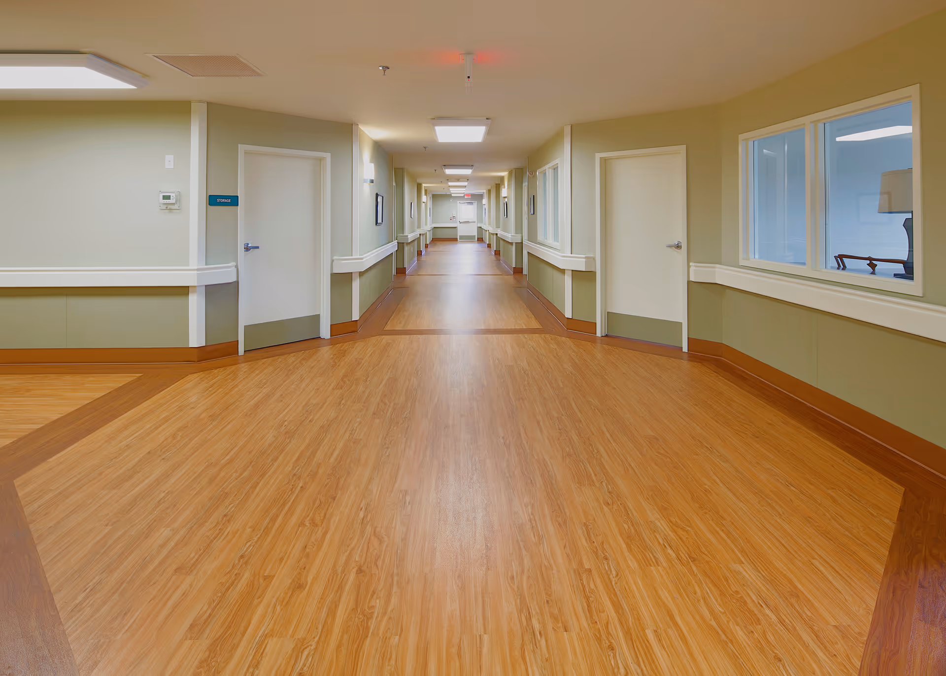 A long, wide hallway in a senior living facility with light green walls, wooden flooring, white doors, and handrails along both sides. The hallway is well-lit with ceiling lights and has windows on the right side showing a room with furniture inside.