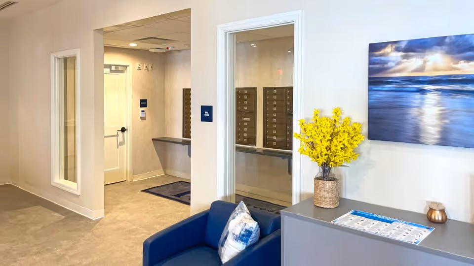 Interior view of a senior living facility lobby area with a blue armchair, a side table holding a vase with yellow flowers, a wall-mounted painting of a seascape, and a hallway leading to a mail room with mailboxes visible through a glass window.
