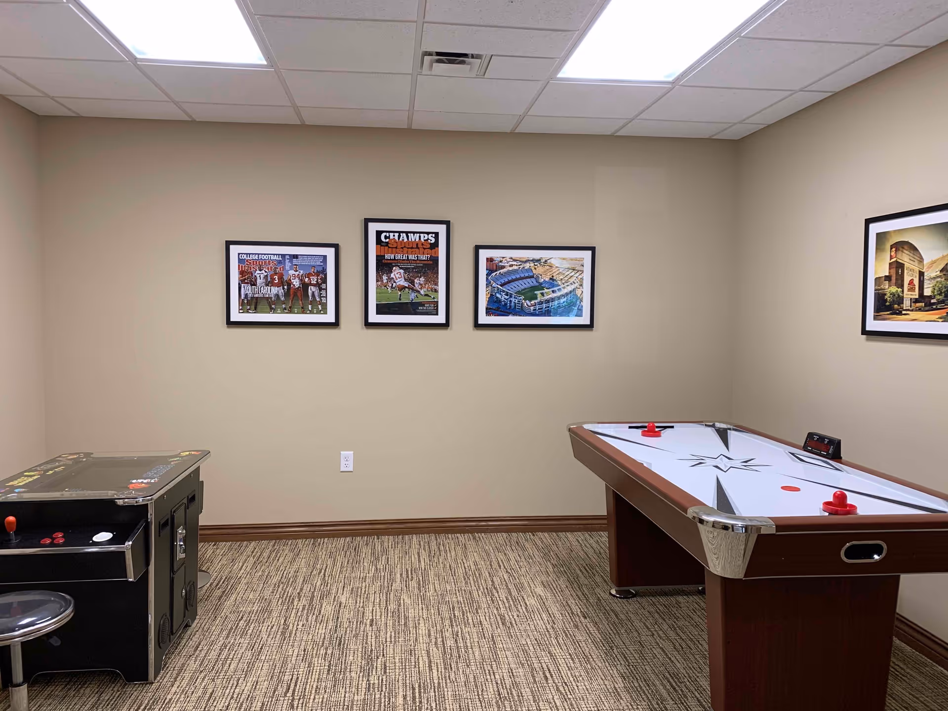 A game room with an air hockey table on the right and a vintage arcade game on the left. The walls are beige with four framed pictures hanging, including sports and stadium images. The floor is carpeted and the ceiling has white tiles with fluorescent lighting.