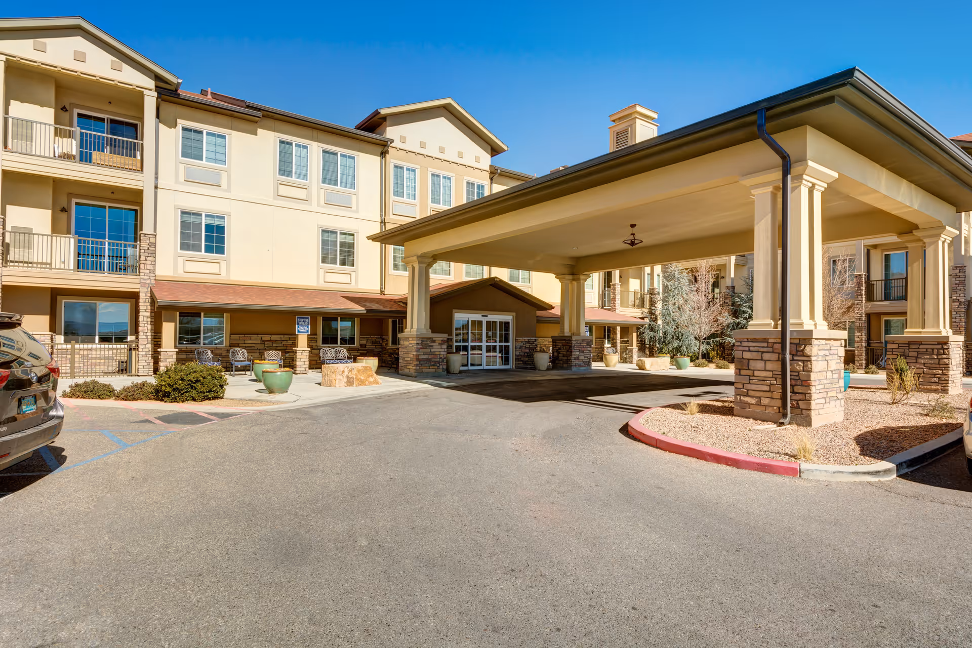 Front entrance of a three-story beige senior living building with a covered porte-cochère, stone columns, balconies, and parked cars.