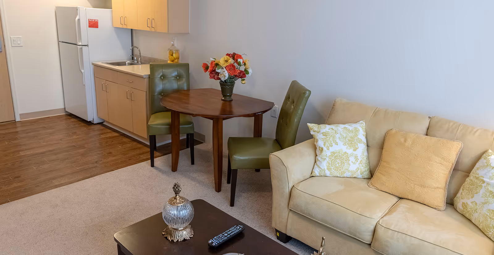 Cozy living area featuring a beige sofa, round wooden dining table with green chairs, and a compact kitchenette with refrigerator and sink.
