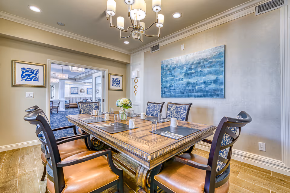 A well-lit dining room with a wooden table set for six people, featuring placemats, glasses, and a vase with flowers in the center. The room has elegant chairs with patterned upholstery, a chandelier overhead, framed artwork on the walls, and an open doorway leading to another dining area with additional tables and chairs.