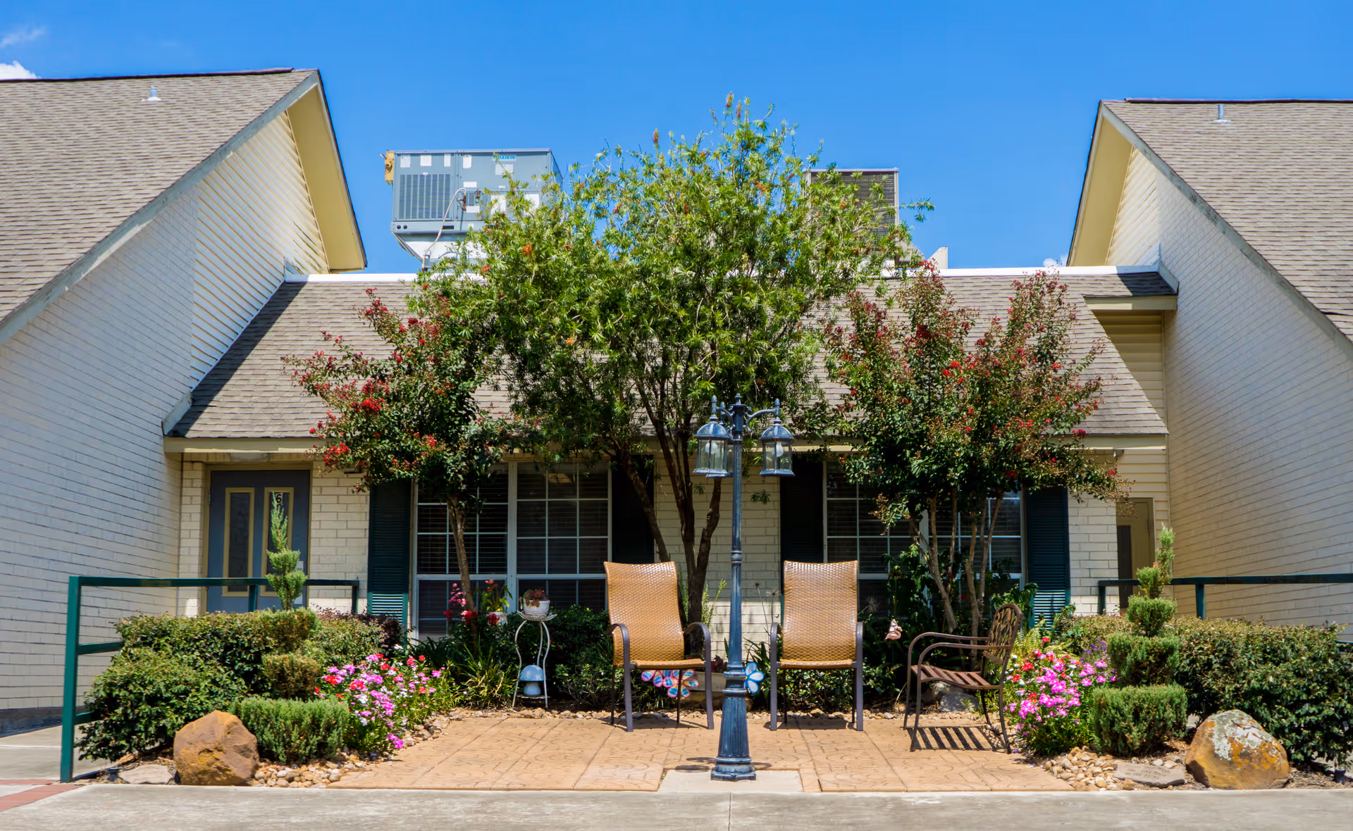 Outdoor seating area in front of a building with two wicker chairs and one metal chair arranged around a black lamppost. The area is surrounded by green bushes, flowering plants, and trees. The building has light-colored brick walls, blue shutters, and a sloped roof with HVAC units visible on top.