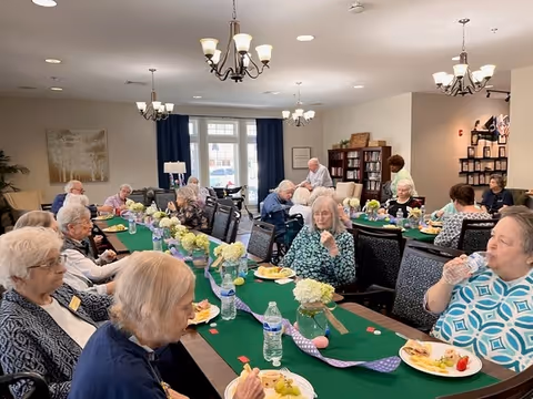 A group of elderly people sitting around a long dining table in a well-lit room, eating and drinking. The table is decorated with green tablecloths, white flower centerpieces, and water bottles. The room has chandeliers, bookshelves, and large windows with curtains.