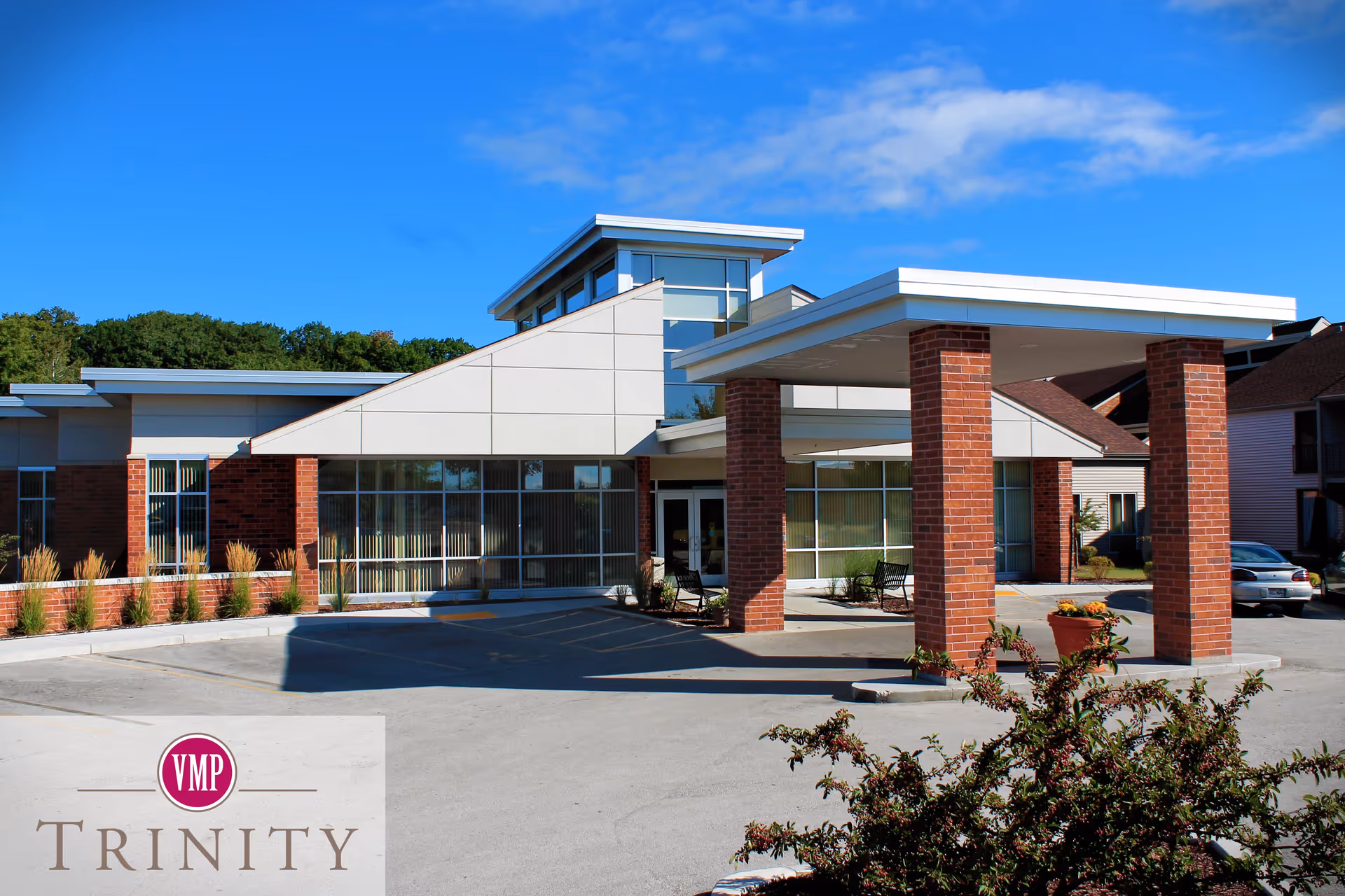 Exterior view of a modern senior living facility building with a covered entrance supported by brick columns, large windows, and a clear blue sky in the background. There are some plants and a parking area in front of the building.