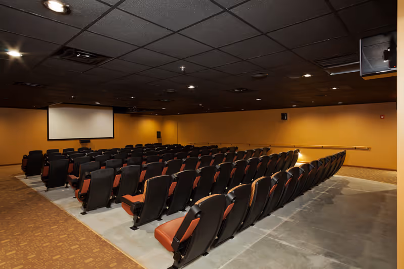 A large room with multiple rows of theater-style seating facing a blank projection screen on the front wall. The room has a dark ceiling with recessed lighting and tan-colored walls. The floor is a combination of carpet and concrete.
