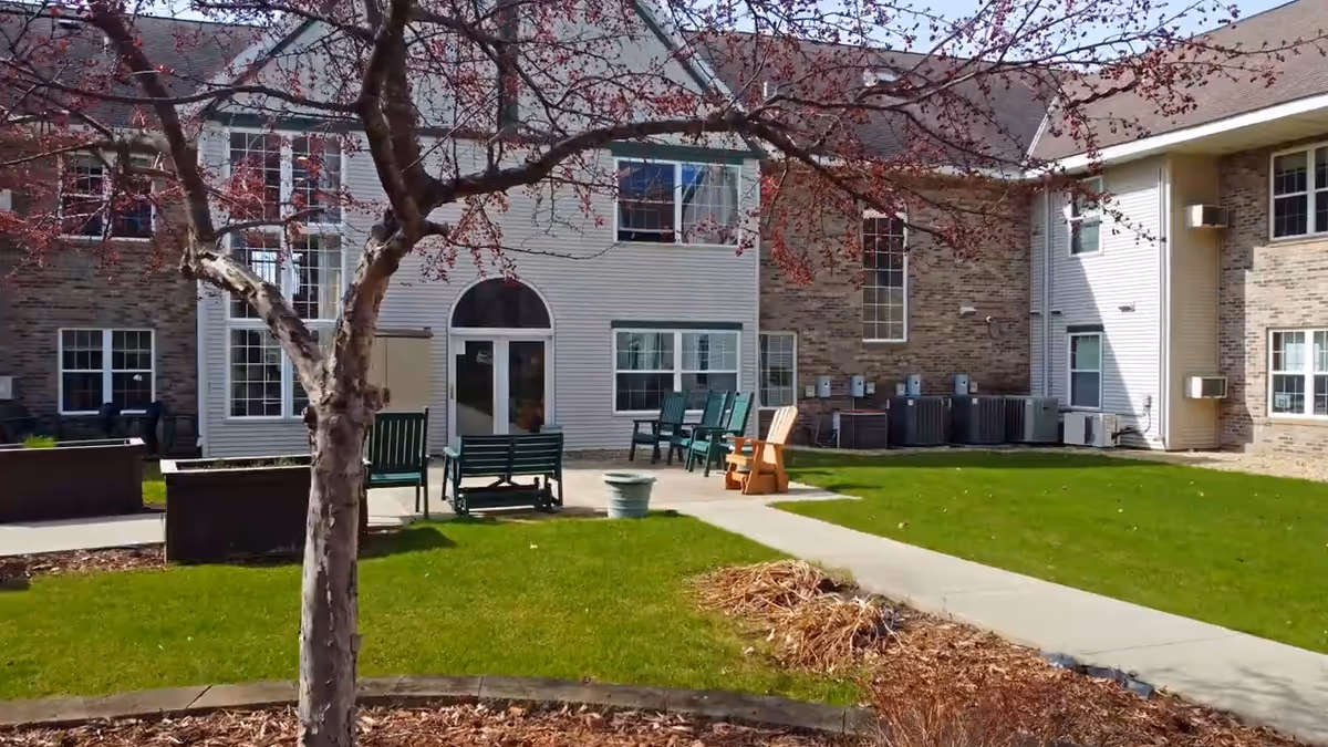 Outdoor courtyard area of a senior living facility with a tree in the foreground, green grass, a concrete walkway, and several chairs and benches arranged on a patio in front of a two-story building with brick and siding exterior.