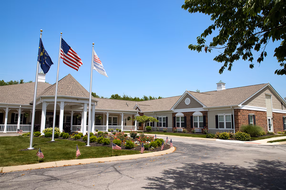 Exterior view of The Commons at Honey Creek senior living facility on a clear sunny day. The building features a covered entrance with white columns and a porch. Three flagpoles display the Indiana state flag, the United States flag, and a flag with the American Communities logo. The surrounding area includes a well-maintained lawn, flower beds, and small American flags along the curb.