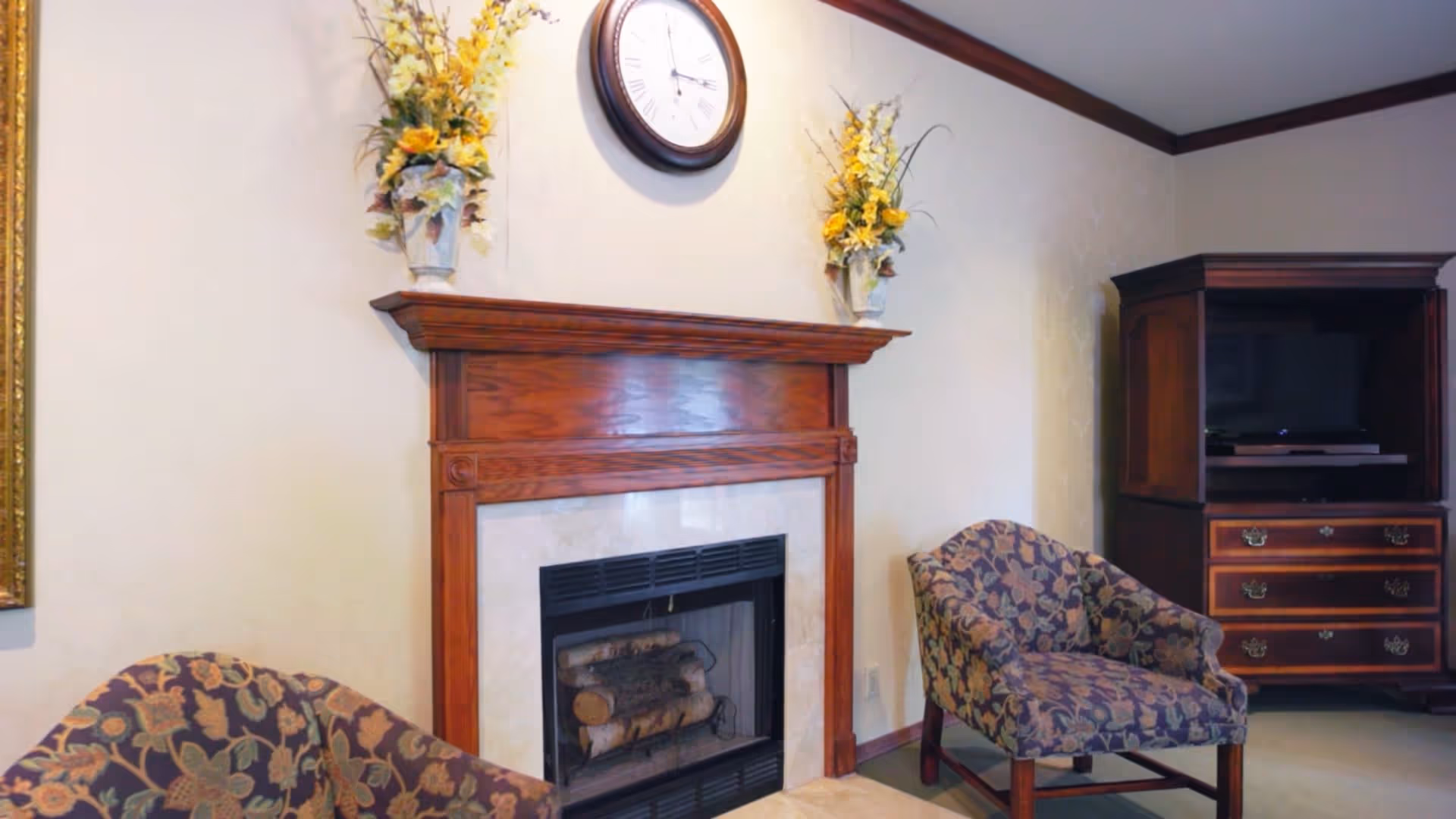 A cozy living room area featuring a wooden fireplace with logs inside, a round wall clock above it, two floral-patterned armchairs, and a wooden cabinet with a television inside.