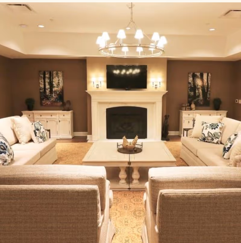 A cozy living room with two beige sofas and two beige armchairs arranged around a white coffee table. The room features a white fireplace with a flat-screen TV mounted above it, flanked by two wall sconces. There are two white cabinets on either side of the fireplace, each decorated with plants and artwork. A large circular chandelier hangs from the ceiling, illuminating the space.