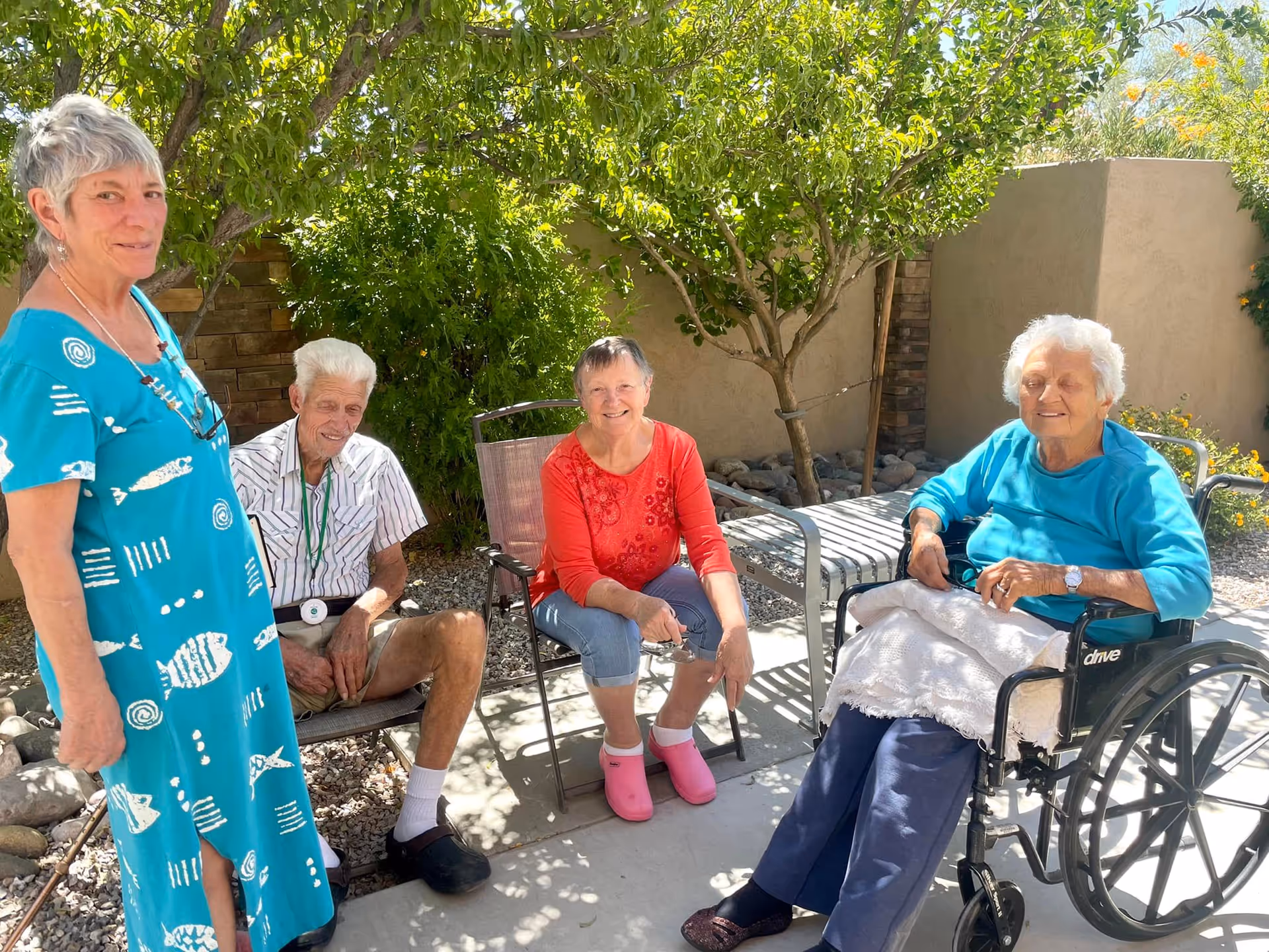 Four elderly individuals sitting and standing outdoors in a shaded garden area. One woman in a blue dress stands on the left, an elderly man in a striped shirt and shorts sits on a chair, a woman in a red shirt and pink shoes sits on another chair, and an elderly woman in a blue top sits in a wheelchair with a white blanket on her lap. Green trees and plants surround them, and a beige wall is in the background.