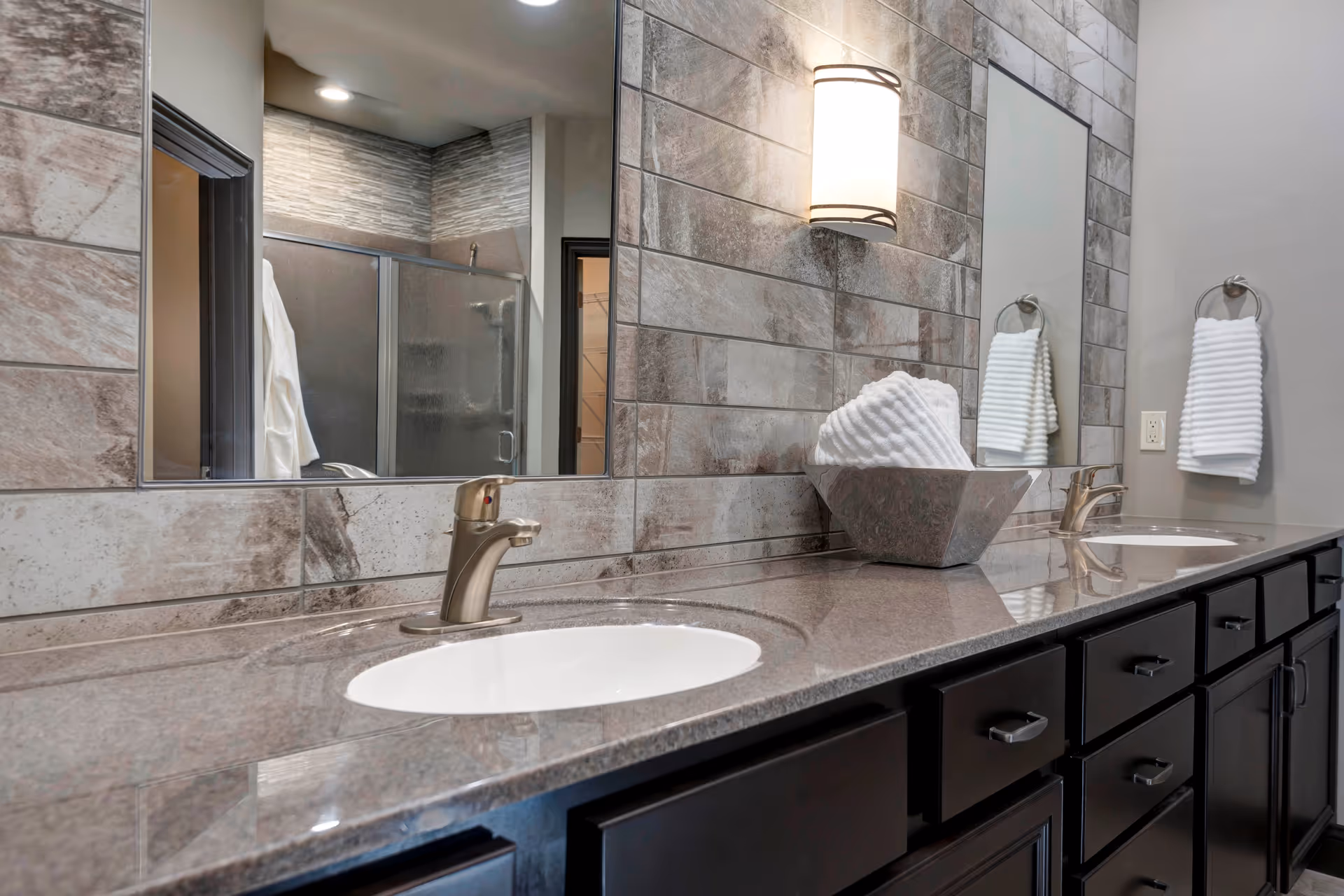 Modern bathroom with a double sink vanity featuring dark wood cabinets and a polished stone countertop. Above the sinks is a large mirror with a wall-mounted light fixture. The wall is tiled with rectangular stone tiles. A towel ring with a white towel is mounted on the wall, and a decorative bowl with folded white towels sits on the countertop. In the reflection, a glass-enclosed shower and a white robe hanging on the door are visible.